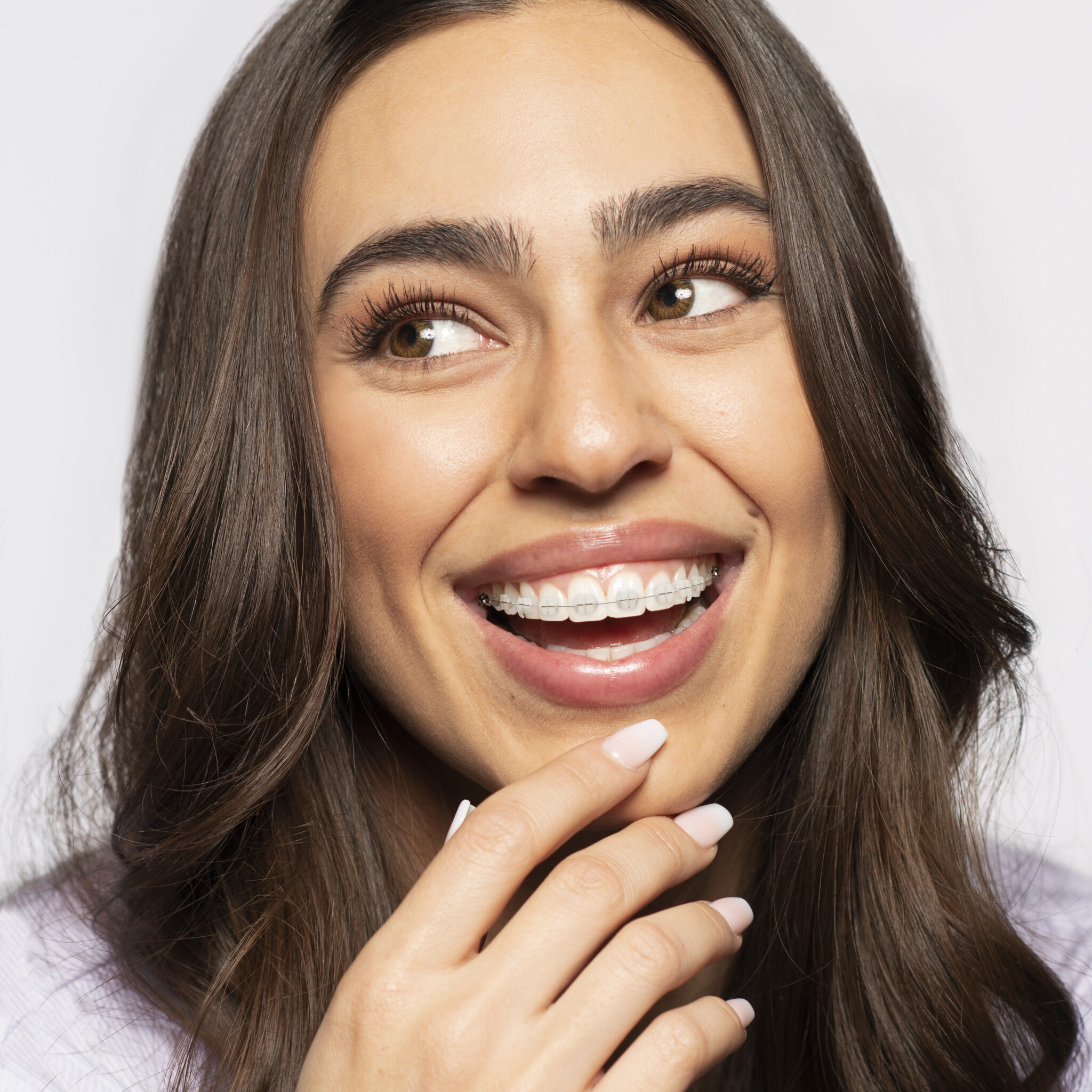 Young woman with long brown hair smiling and showing teeth with braces, holding her chin with one hand, showcasing the expert care from Redmond Ridge Orthodontics.