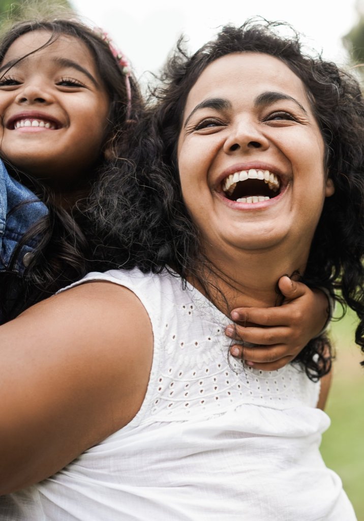 A woman with curly hair and a white shirt smiles broadly while giving a piggyback ride to a young girl in a denim jacket. Both are outdoors, near Redmond Ridge Orthodontics, and appear joyful.