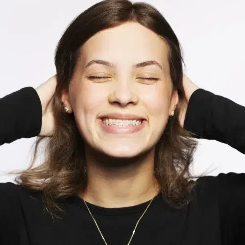 A person with brown hair and a black shirt, representing Redmond Ridge Orthodontics, smiles with eyes closed and hands behind their head against a plain white background.
