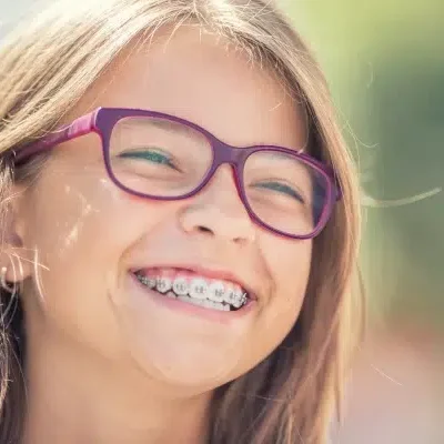 A young girl with long hair, wearing purple glasses and braces from Redmond Ridge Orthodontics, is smiling outdoors.